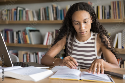 Focused African schoolgirl or student girl studying in college library, reading topics while sit at desk with laptop and textbooks, engaged in personal development, self-education, exams preparations