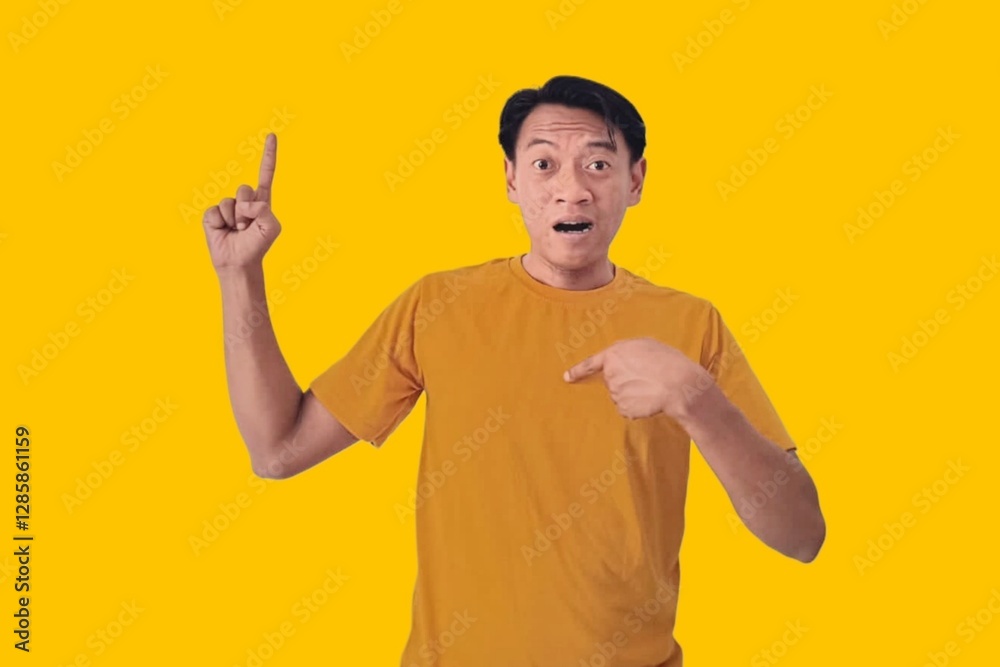 A young Asian man in a yellow shirt looks surprised, pointing enthusiastically at something off-frame against a yellow background. The image captures an expressive moment of amazement A young Asian man in a yellow shirt looks surprised, pointing enthusiastically at something off-frame against a yellow background. The image captures an expressive moment of amazement