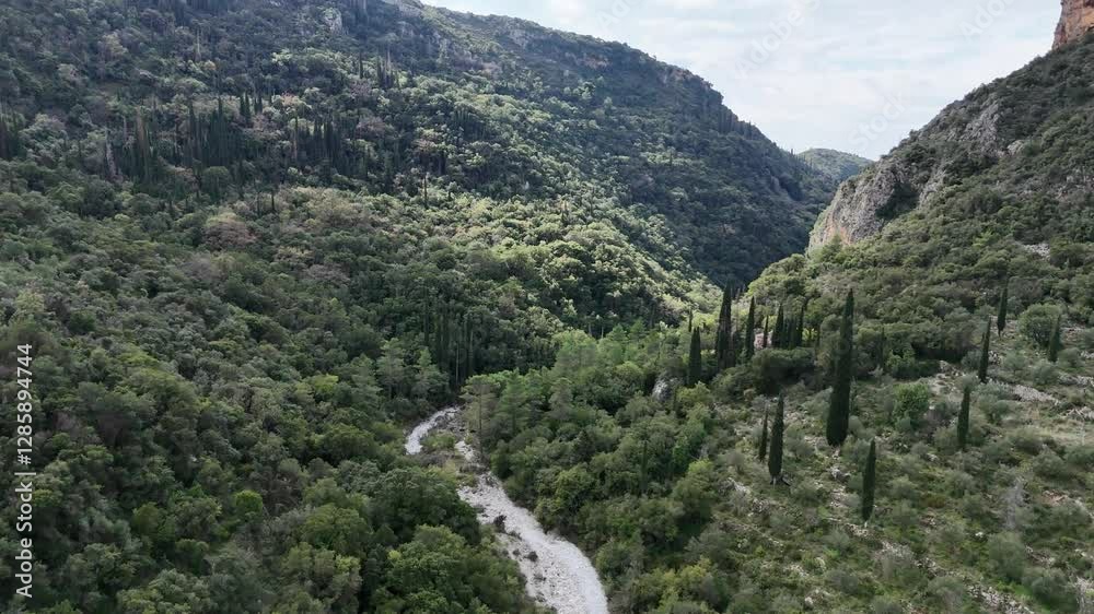 Aerial drone flight through Vyros Gorge near Kardamyli, Greece, on a sunny winter day. The gorge features lush green vegetation, striking red rock formations, and surrounding mountains. Aerial drone flight through Vyros Gorge near Kardamyli, Greece, on a sunny winter day. The gorge features lush green vegetation, striking red rock formations, and surrounding mountains.