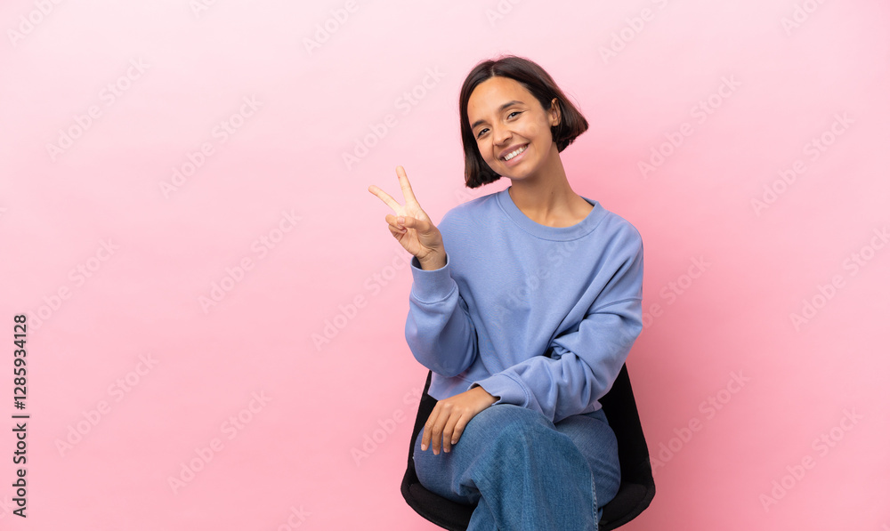 Young mixed race woman sitting on a chair isolated on pink background smiling and showing victory sign Young mixed race woman sitting on a chair isolated on pink background smiling and showing victory sign