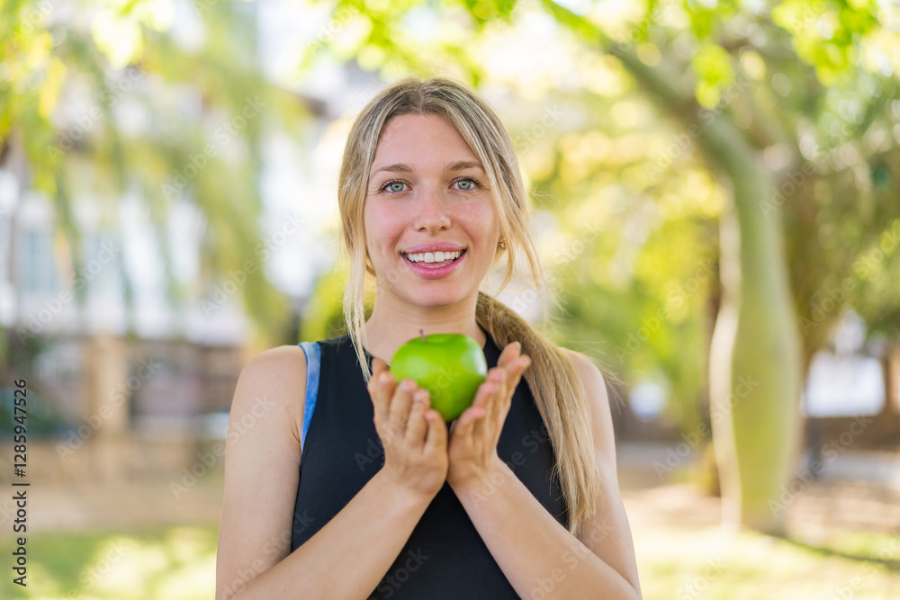 Young blonde woman at outdoors holding an apple with happy expression Young blonde woman at outdoors holding an apple with happy expression