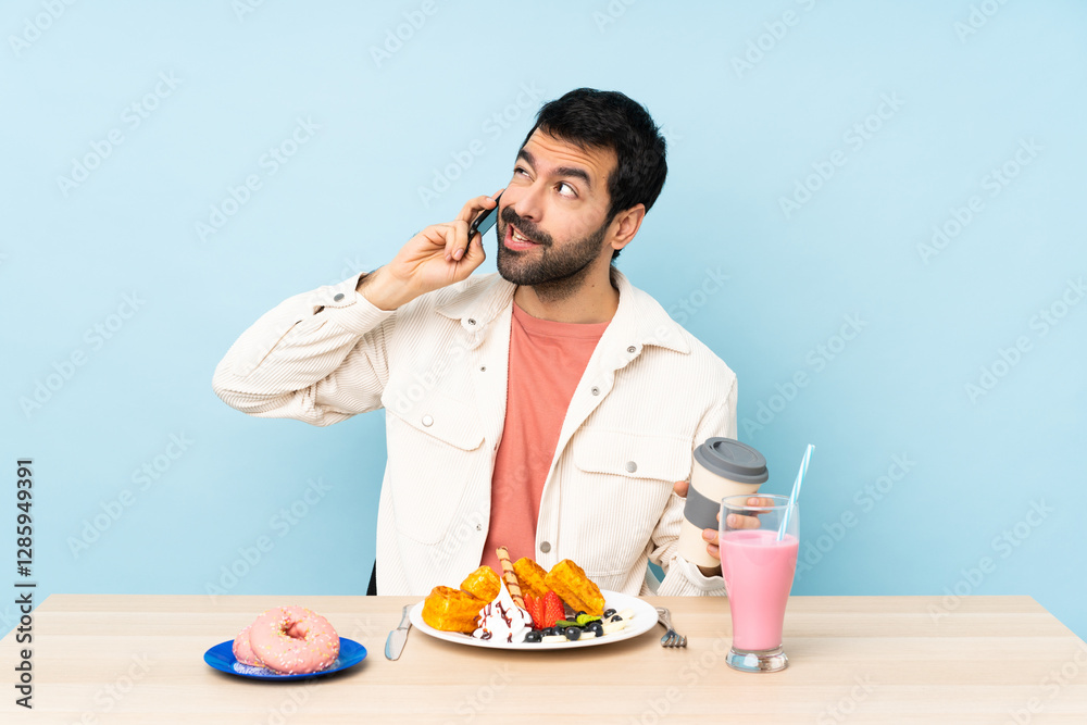 Man at a table having breakfast waffles and a milkshake holding coffee to take away and a mobile