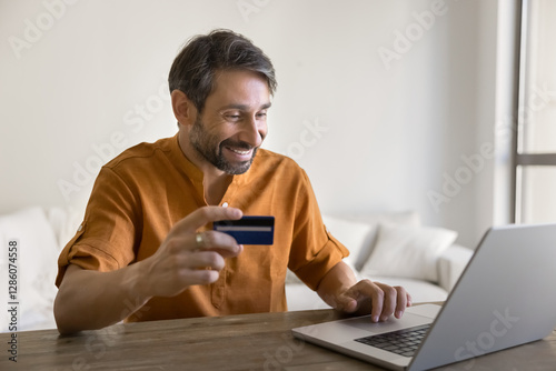 Smiling Portuguese 40s man doing online shopping at home, sit at table with laptop, hold debit or credit card, buying gifts, spend money, making quick transactions. Cashless payments via marketplace