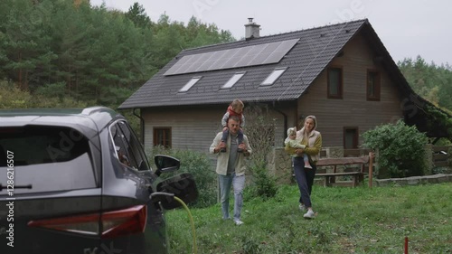 Parents with kids walking to their electric car parked in front of house. House with rooftop solar panels and own EV home charging station.