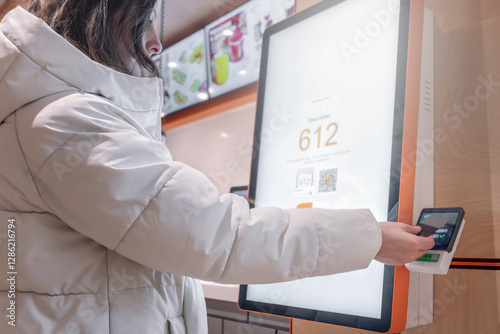 A woman dressed in a stylish white coat is making a convenient mobile payment at a modern selfservice kiosk A white woman in a jacket pays for an order with a bank card in a restaurant through a