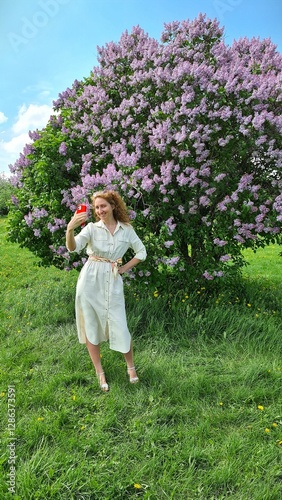 a girl takes a selfie in the park on a summer sunny day against the background of a blooming siren tree and green grass and blue sky