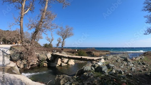  A scenic rustic landscape on Samothrace Island, Greece, featuring a small wooden bridge over a stream leading to the sea. The area is surrounded by rocks, dry trees, and a beautiful coastal view. Goa