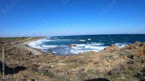 waves crash against a rugged, rocky shoreline under a clear blue sky. The white foam contrasts with the deep blue sea, creating a dynamic coastal scene.