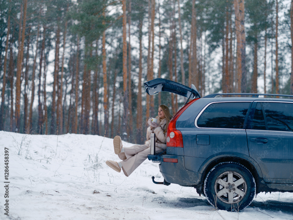 Fototapeta premium Happy young woman sitting in opened car trunk and drinking hot tea or coffee in snowy forest.