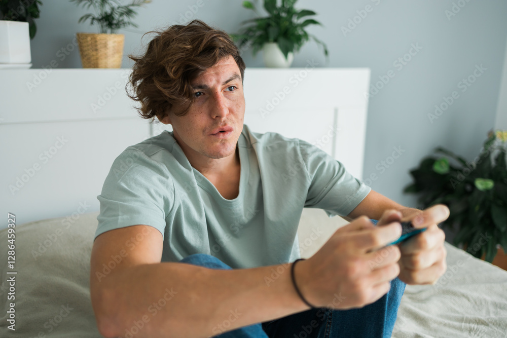 Handsome excited gamer sitting on a bed and playing video games on a console. Wireless controller and online game technologies concept Handsome excited gamer sitting on a bed and playing video games on a console. Wireless controller and online game technologies concept