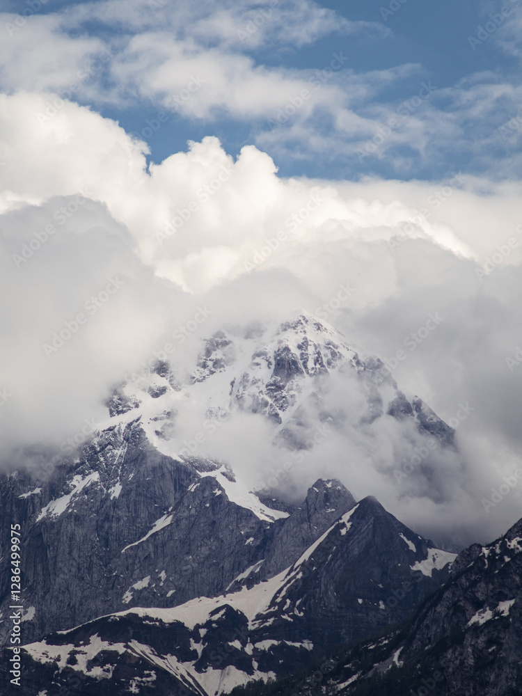 A majestic snow-covered mountain peak partially hidden by thick clouds in the Friuli Venezia Giulia region, Italy. The rugged rock formations contrast with the soft, billowing clouds. A majestic snow-covered mountain peak partially hidden by thick clouds in the Friuli Venezia Giulia region, Italy. The rugged rock formations contrast with the soft, billowing clouds.