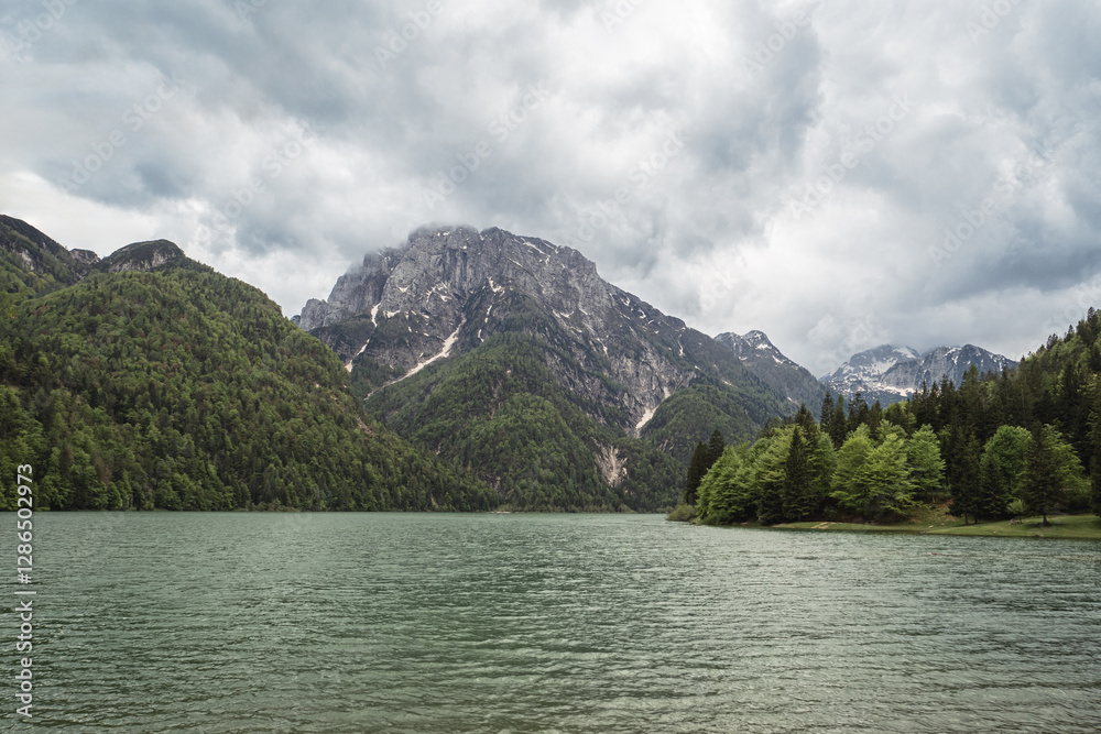 A tranquil view of Lago del Predil in Italy, surrounded by lush green mountains and towering rocky peaks. The calm turquoise waters reflect the dramatic cloudy sky above. A tranquil view of Lago del Predil in Italy, surrounded by lush green mountains and towering rocky peaks. The calm turquoise waters reflect the dramatic cloudy sky above.