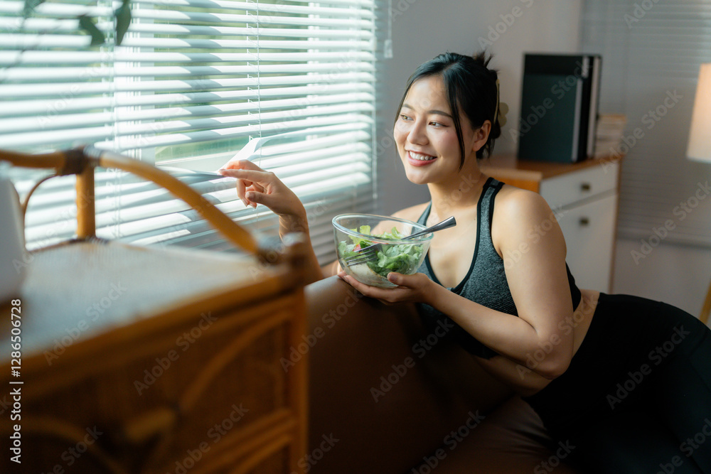 Young woman in sportswear sitting on living room sofa, consuming healthy salad from glass bowl while pulling blinds open for natural lighting Young woman in sportswear sitting on living room sofa, consuming healthy salad from glass bowl while pulling blinds open for natural lighting