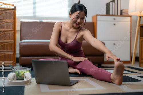 Smiling young Asian female doing yoga stretch near laptop, following virtual fitness class, embracing wellness routine and personal health goals