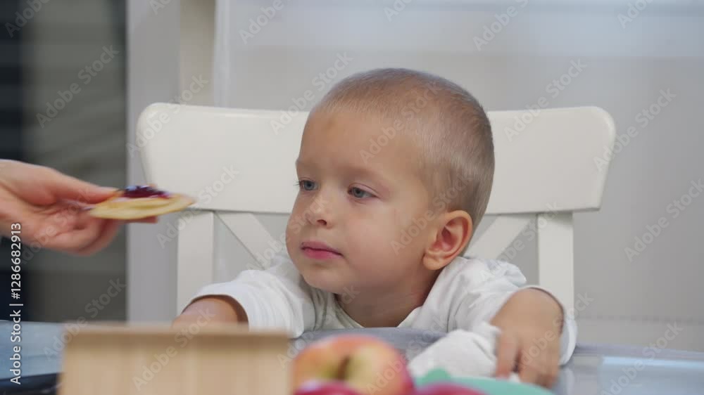 Mother treating her little son to pancakes with jam. Woman and child sitting at the dining table and eating pancakes. Mother treating her little son to pancakes with jam. Woman and child sitting at the dining table and eating pancakes.