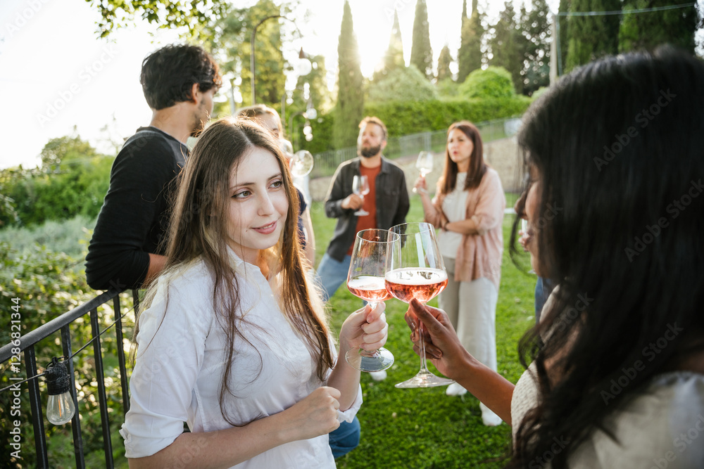 Fototapeta premium Group of friends celebrating outdoors drinking rosé wine on a sunny day - A couple of women look at each other and toasting with two glasses