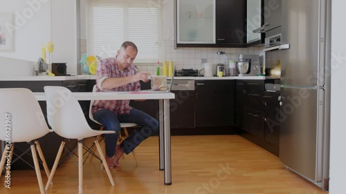 A man sits at a kitchen table using his laptop for work. His children are playing nearby, creating a lively atmosphere in the cozy home environment during the day.
