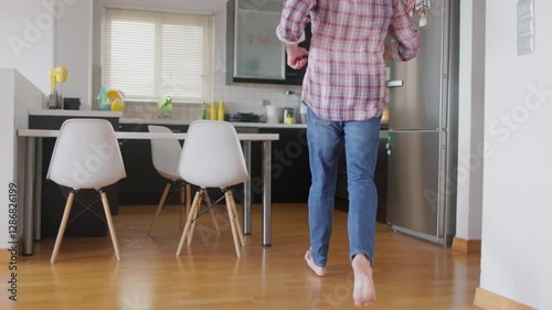 A father is focused on cooking a meal while his children run and play nearby in a cheerful kitchen setting, slow motion. Sunlight brightens the space, creating a warm atmosphere.