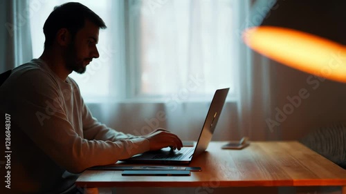 Warm light from lamp creates cozy atmosphere for deep concentration. Man sits at desk, engaged with laptop, surrounded by a serene evening setting