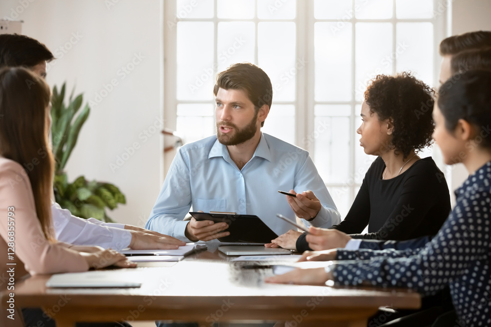 Fototapeta premium Serious skilled teammates gathered together at conference table for decision making, planning strategy, forecasting. Millennial team leader speaking during briefing event with corporate staff members