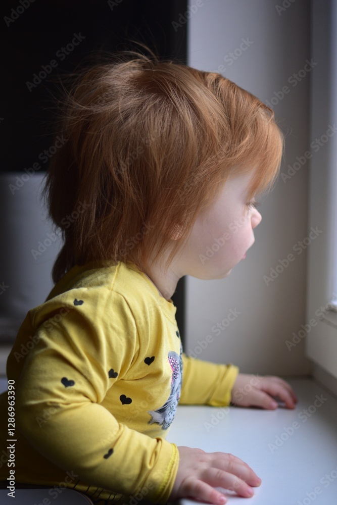 Fototapeta premium Red messy hair toddler girl curiously looking through the window. Daughter impatiently waiting parents.