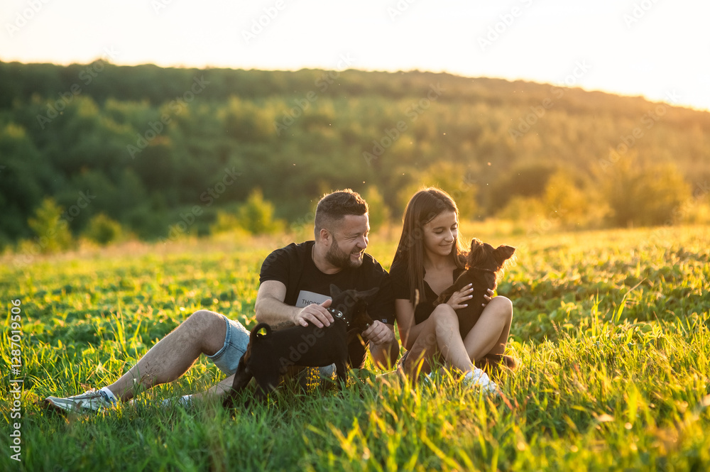 Fototapeta premium A cute little dog stands on its hind legs. A happy family plays together in the park with a pinscher dog. The couple is dressed in blue shorts and black T-shirts.