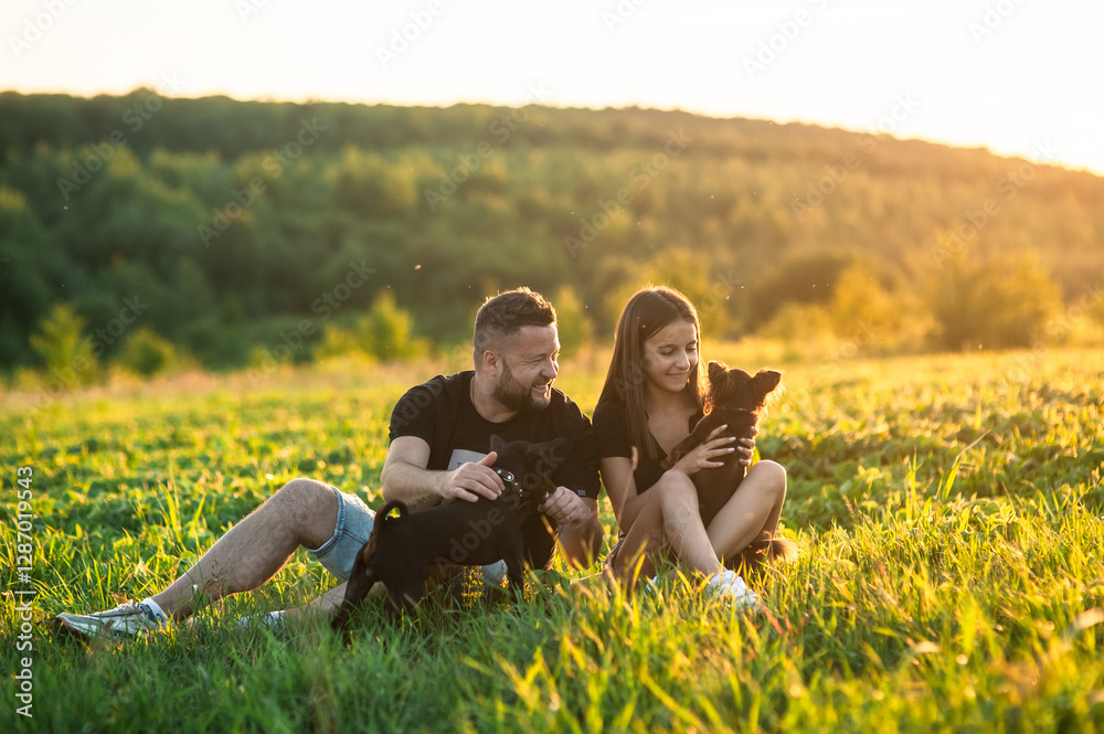The couple is dressed in blue shorts and black T-shirts.Сouple is playing with a kind black domestic dog in the forest, on a summer day. The couple is dressed in blue shorts and black T-shirts.Сouple is playing with a kind black domestic dog in the forest, on a summer day.