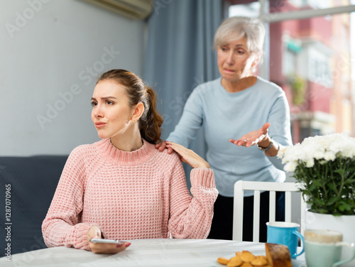 Portrait of offended young woman sitting at home table, listening to reproaches from her aged mother. photo