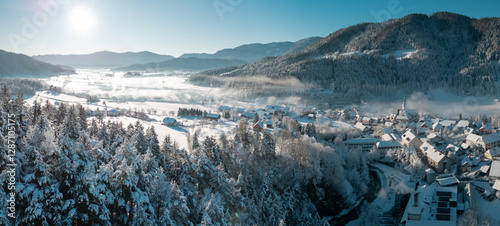 Obraz na płótnie Beautiful village of Ljubno ob Savinji in cold winter morning. Visible haze from the river, sun rising up and smoke coming out of smokestacks. w salonie