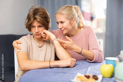 Portrait of offended teenager is soothed by caring mom while sitting at table in cozy dining room.. photo