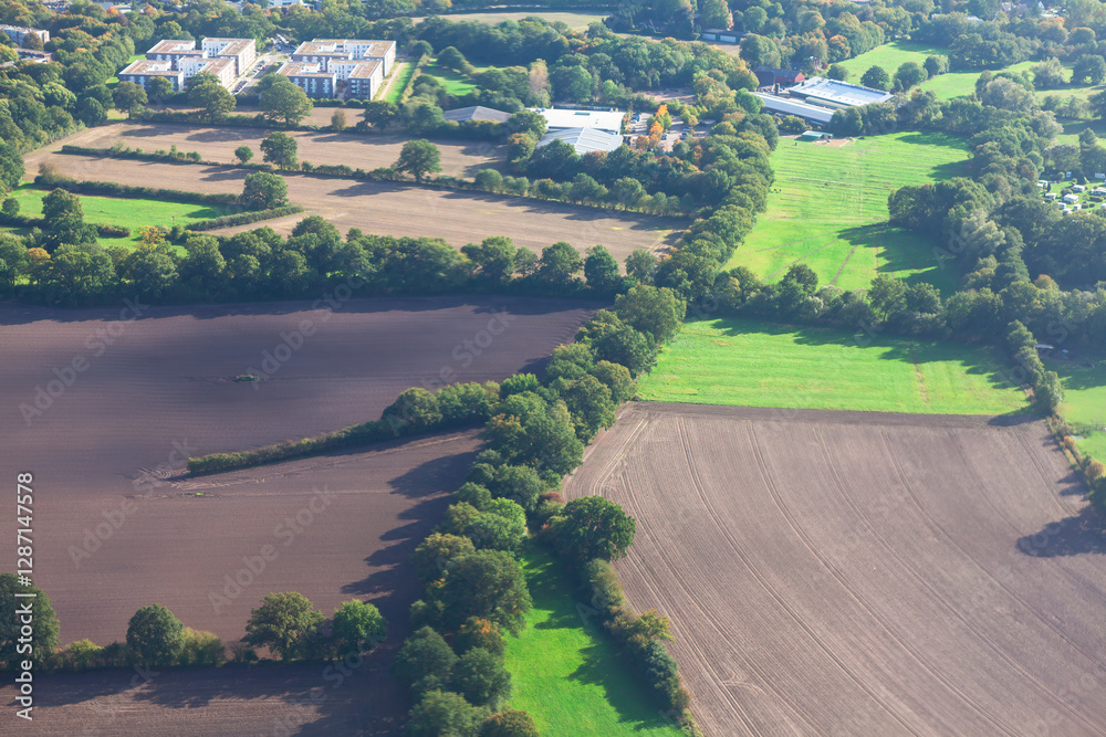 Aerial view of a rural landscape with fields, trees, and some buildings in the background. Fields are divided by rows of trees, and the buildings appear to be part of a small settlement or farm