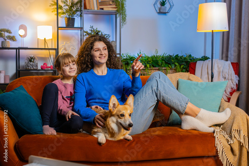 Surprised mother and daughter child girl sitting beside corgi dog watching TV in living room at home. Happy family in casual clothes watching favorite TV show sits on sofa in apartment during weekend.