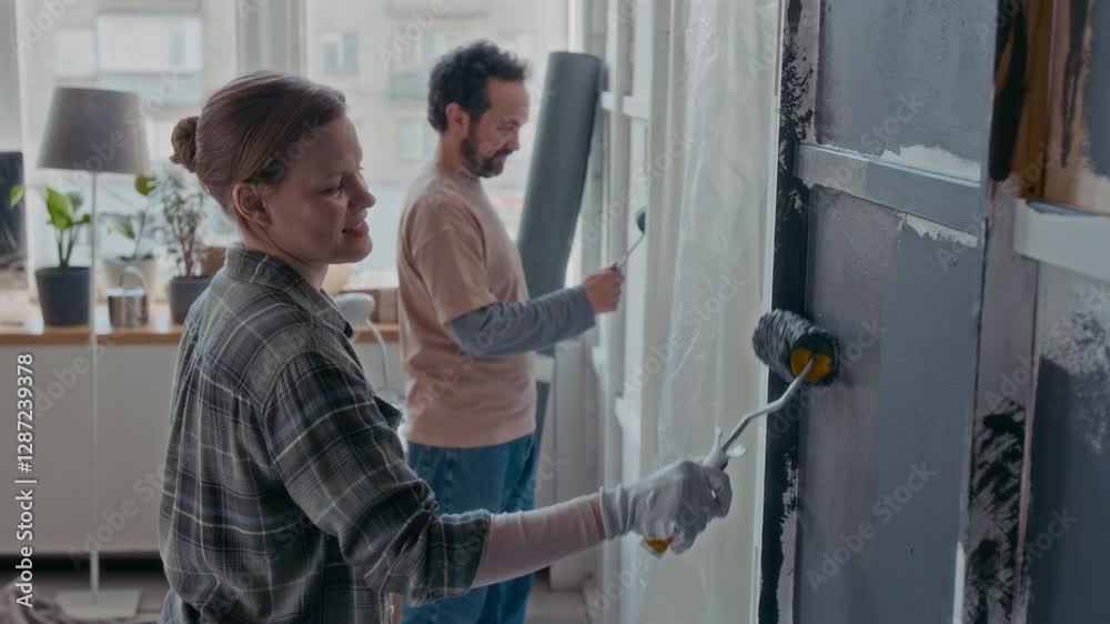 Side view of young woman with paintroller applying grey paint on wall during renovation while standing against her husband working with another part of domestic room