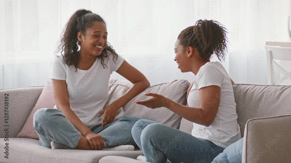 Two African American sisters relaxing on the couch in a cozy home setting. They are engaged in a warm conversation, showcasing their close bond and affection for each other.