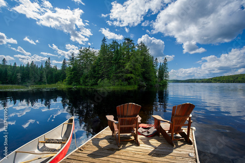 Two Adirondack chairs on a wooden dock overlook the blue waters of a Muskoka lake. A life jacket and paddles are visible, while a red canoe is tied to the dock.