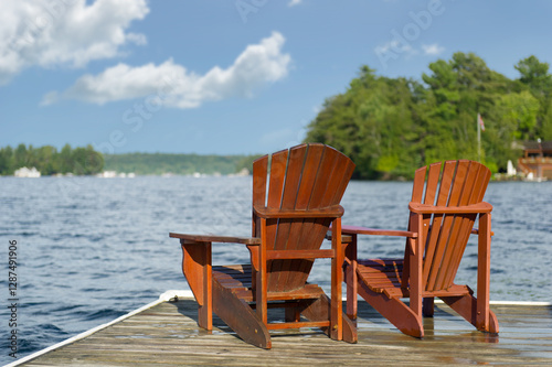 Two Adirondack chairs sit on a wooden dock overlooking a lake. The water is slightly wavy, with cottages nestled among green trees across the shore.