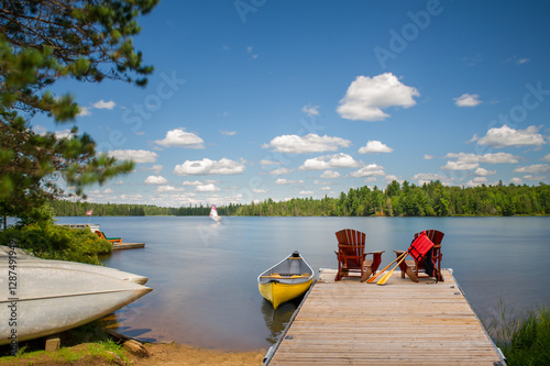 Two Adirondack chairs on a wooden dock overlook the calm blue waters of a Muskoka lake. A life jacket and paddles hang from one chair. A yellow canoe is tied to the dock, with others on the shoreline.