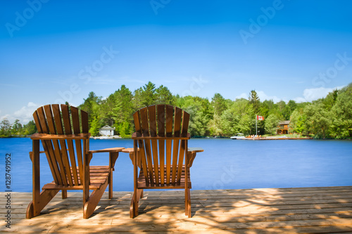 Two Muskoka chairs sit on a wooden dock facing a calm lake. Across the water, cottages are nestled among green trees, with a boat dock extending into the lake. A Canadian flag waves in the breeze.