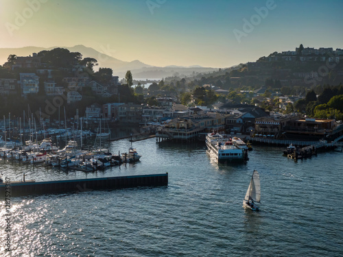Aerial view of a sailboat navigating Tiburon harbor, with docked yachts and a ferry. Coastal town nestled on hillsides. Belvedere Tiburon, San Francisco, California, USA