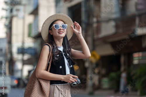 Young woman tourist wearing a stylish hat and sunglasses, holding her hat and camera, happily exploring an asian city, capturing the vibrant culture and urban scenery