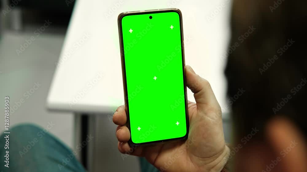 Hand of young man holding smartphone green screen in business office work. Mobile phone vertical mockup. Male person using phone with blank green background display. Chroma key smart phone. Close up.