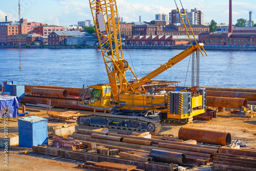 A construction crawler crane on the shore installs supports in the daytime during the construction of a bridge across the river