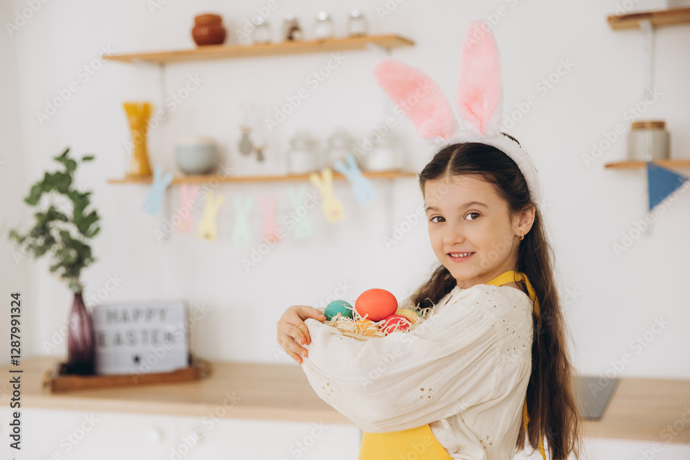 A cute little girl wearing plush bunny ears and apron holding basket full of Easter colorful eggs