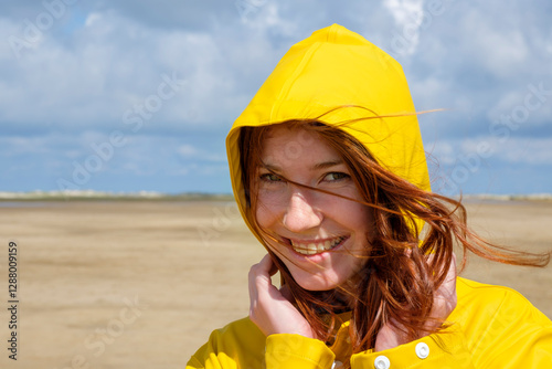 Close-up portrait of carefree redhead teenage girl wearing yellow raincoat at beach against sky on sunny day