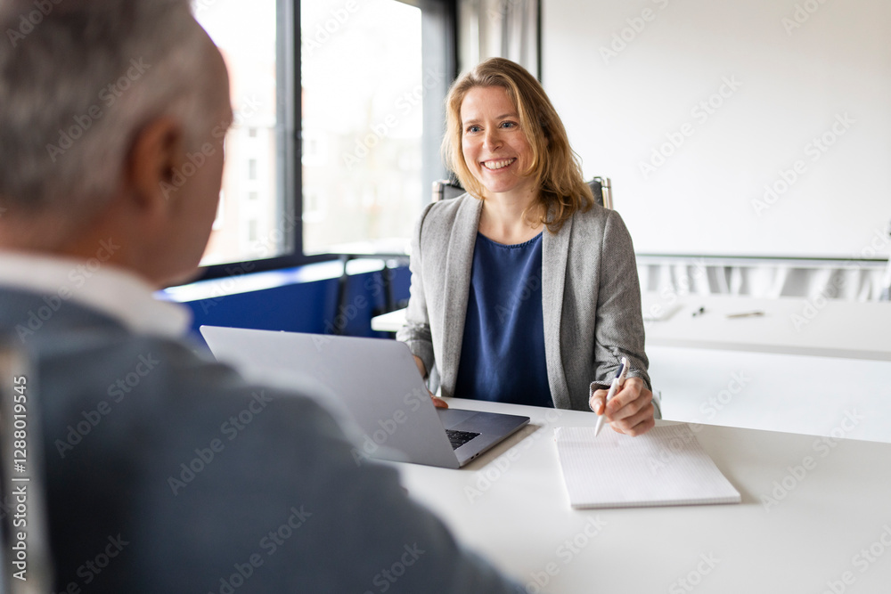 Happy businesswoman sitting near laptop and talking with coworker in office Happy businesswoman sitting near laptop and talking with coworker in office