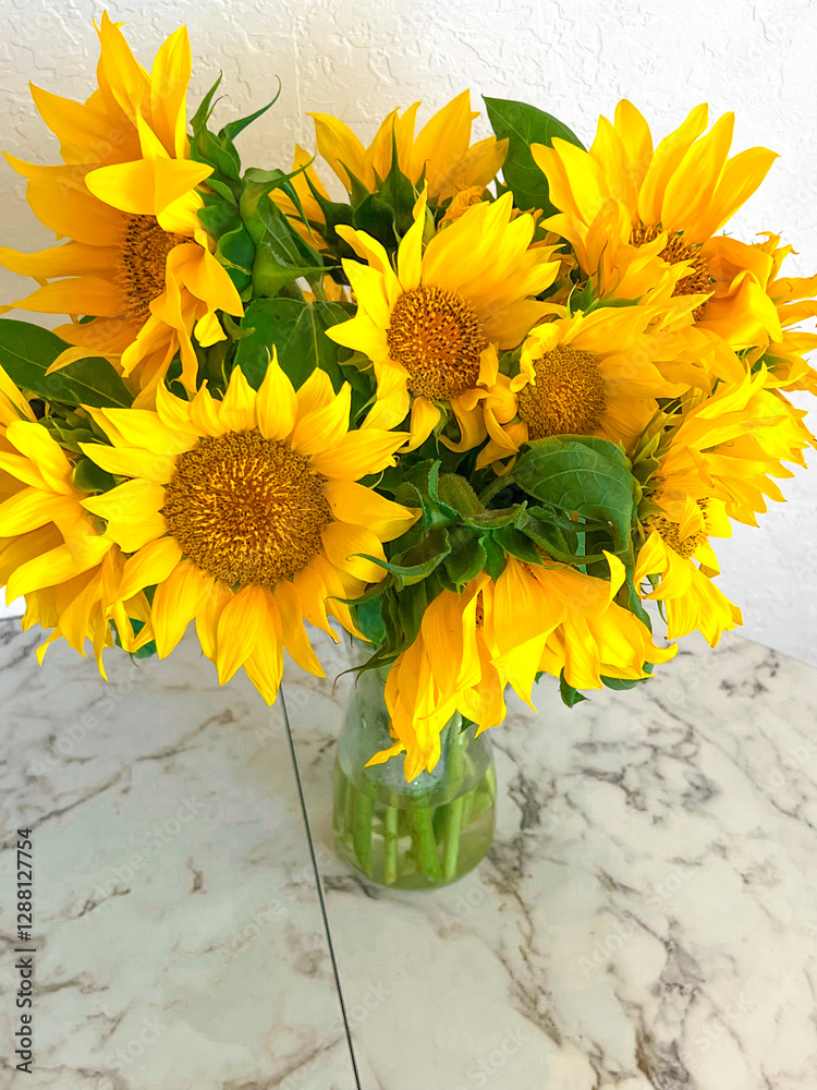 A bouquet of sunflowers stands in a glass vase on a marble table in a white kitchen