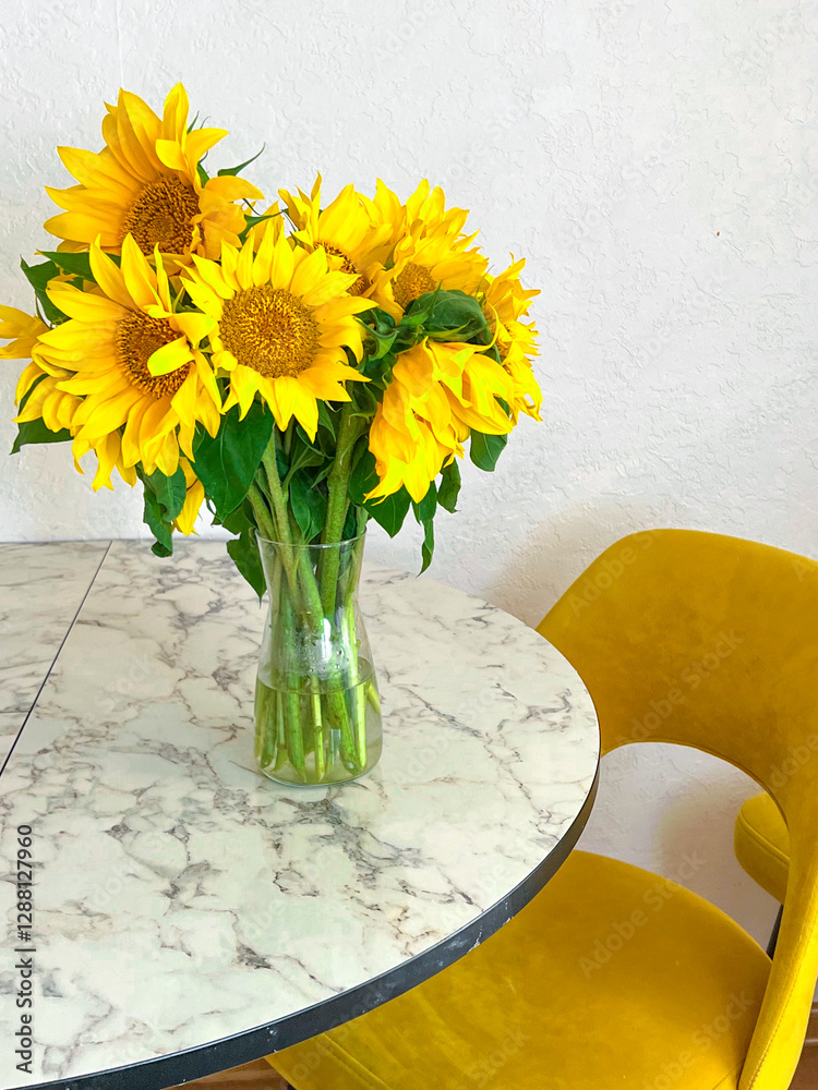 A bouquet of sunflowers stands in a glass vase on a marble table in a white kitchen with yellow armchairs. The kitchen interior is decorated in white tones