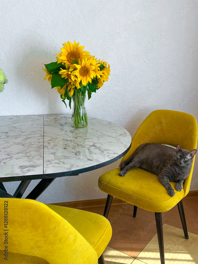 A grey cat is lying on a yellow armchair near a table with a bouquet of sunflowers