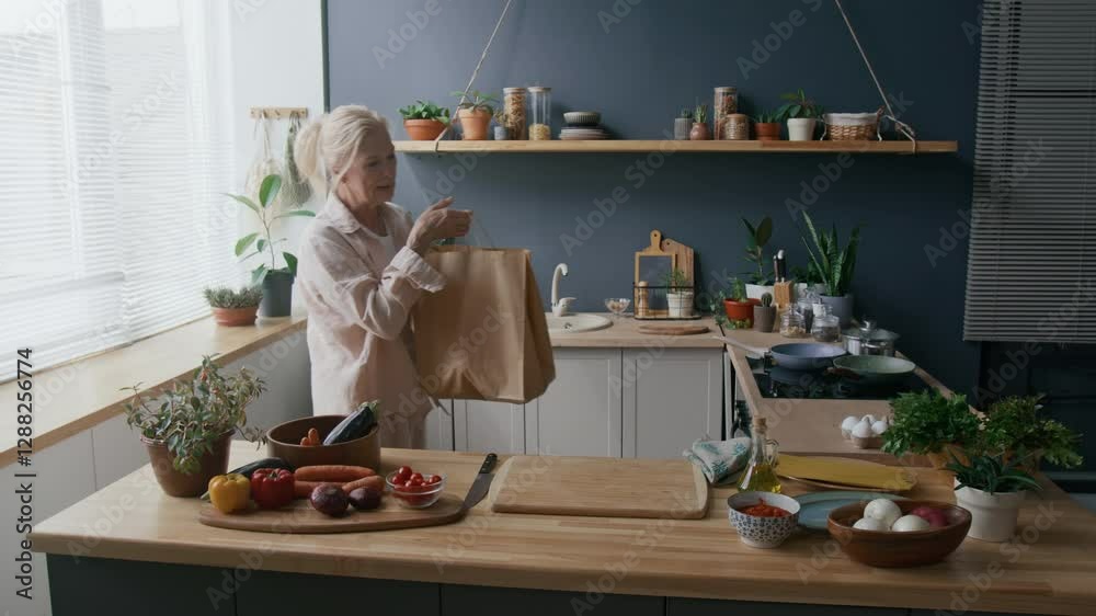 Medium shot of senior Caucasian woman organizing items from brown paper bag in modern kitchen filled with natural light, plants, and fresh produce, creating warm and homely atmosphere