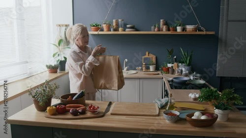 Medium shot of senior Caucasian woman organizing items from brown paper bag in modern kitchen filled with natural light, plants, and fresh produce, creating warm and homely atmosphere